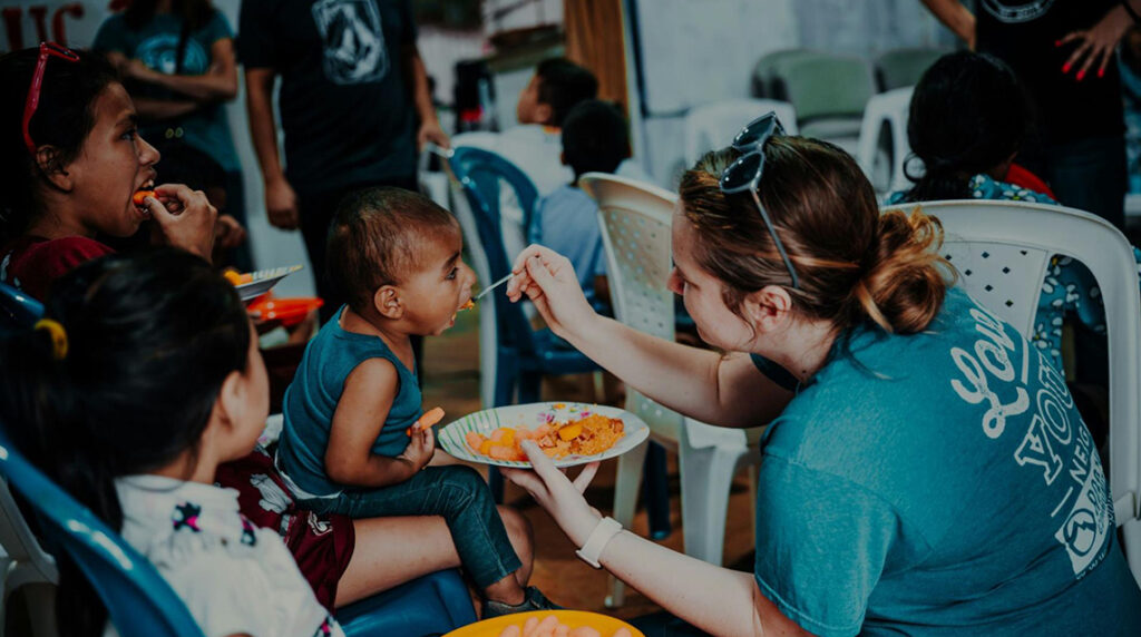 A woman holding a plate of food and kneeling while feeding a child.