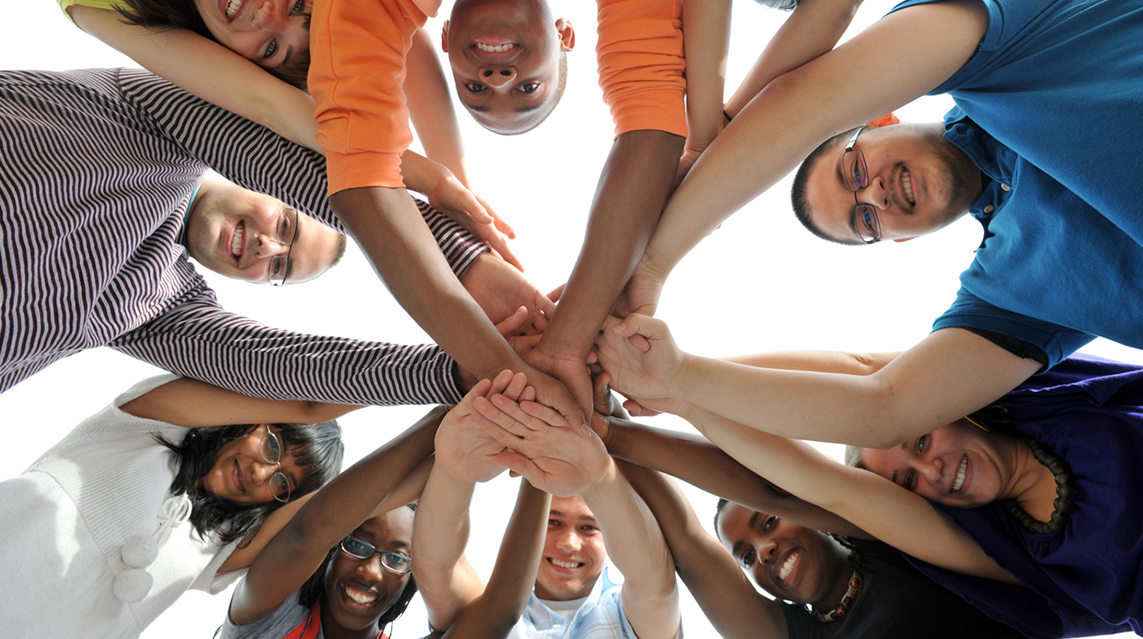 A group of ten young adults who are part of a youth group stand together in a circle and look down, heads meeting in the middle, to smile at the camera.