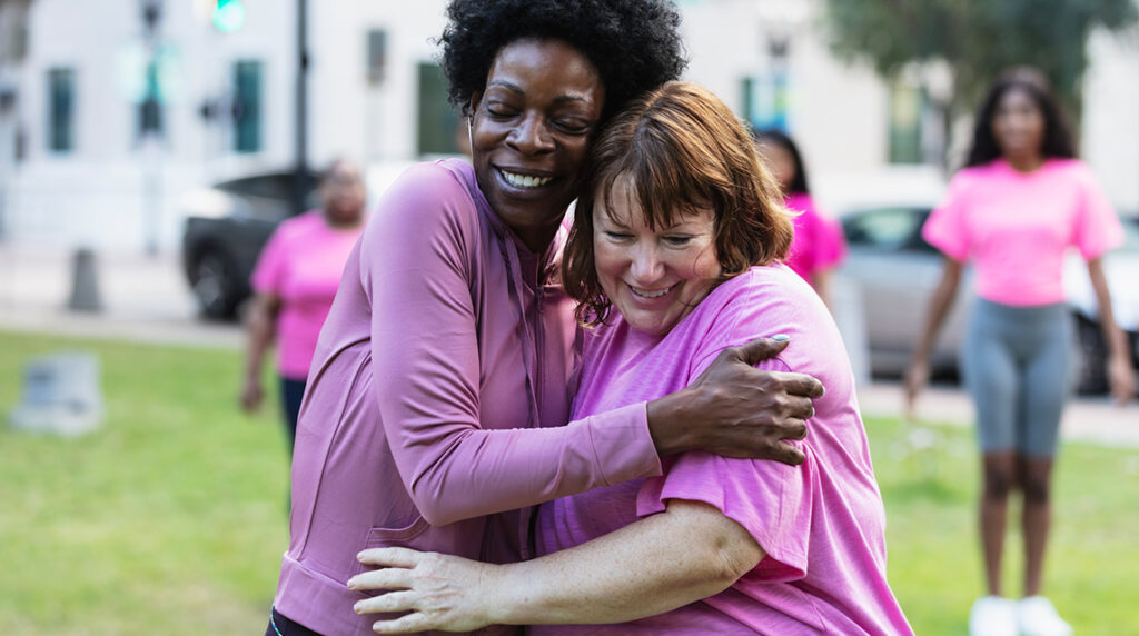 Two multiracial mature women wearing pink hug each other at a breast cancer awareness rally.