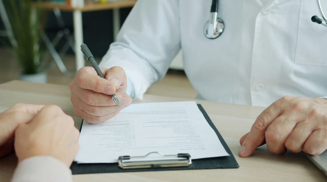 A doctor in a lab coat writing on a clipboard on a desk.