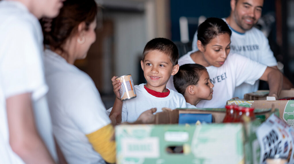 Diverse volunteers pack food boxes in a donation center as a smiling child looks on, reflecting community impact and collaboration in the nonprofit sector.