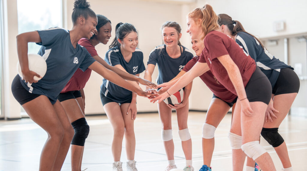 A group of young women huddle together in a circle and place their hands together in the middle to symbolize camaraderie and teamwork.
