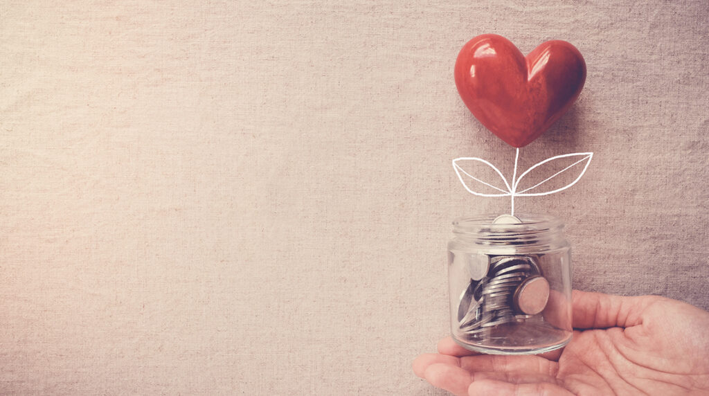 A hand holds a small, clear jar filled with coins with a heart tree sprouting from between them, representing fundraising