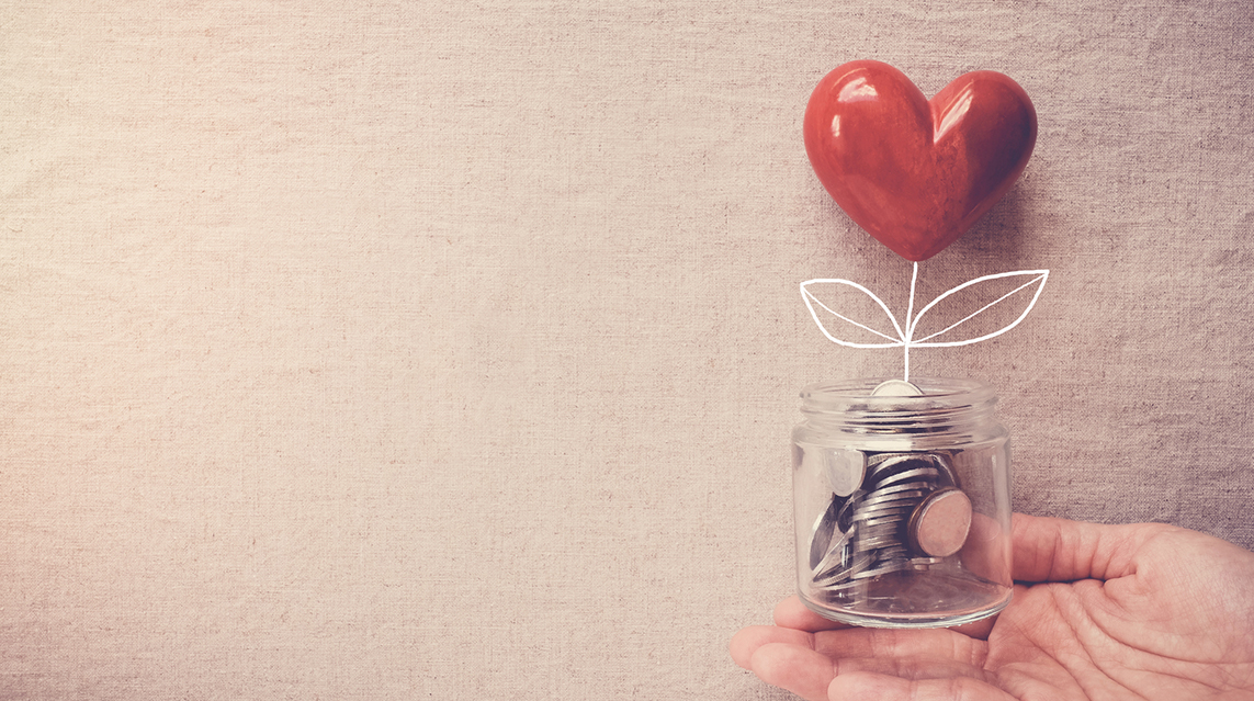 A hand holds a small, clear jar filled with coins with a heart tree sprouting from between them, representing fundraising