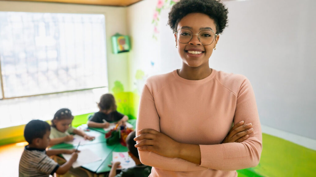 A daycare worker smiling in a classroom while young children work on an activity in the background.