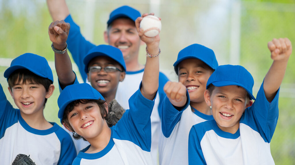 Members of a youth baseball team and their coach stand together and celebrate a win by pumping their fists in the air.