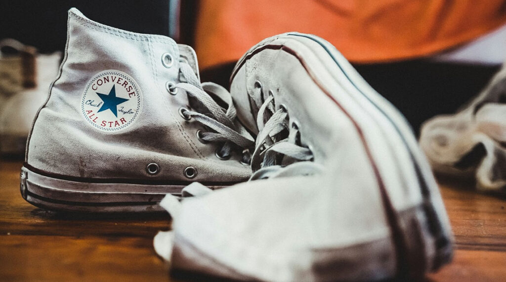 A close-up of an old pair of white Converse sneakers sitting on a wooden floor.