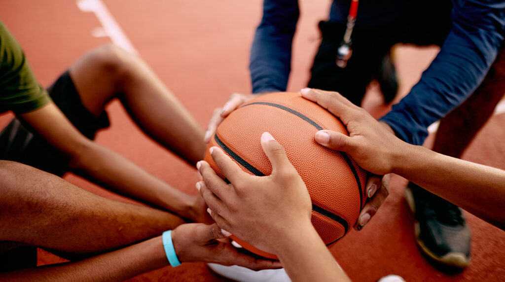 Coach and teen basketball players hold a ball together during practice, showing teamwork that supports successful fundraising ideas for basketball teams.