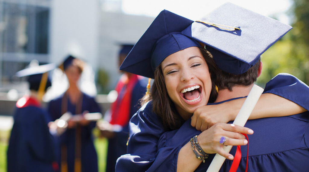 A student smiling and hugging another student, both wearing blue graduation gowns and caps.