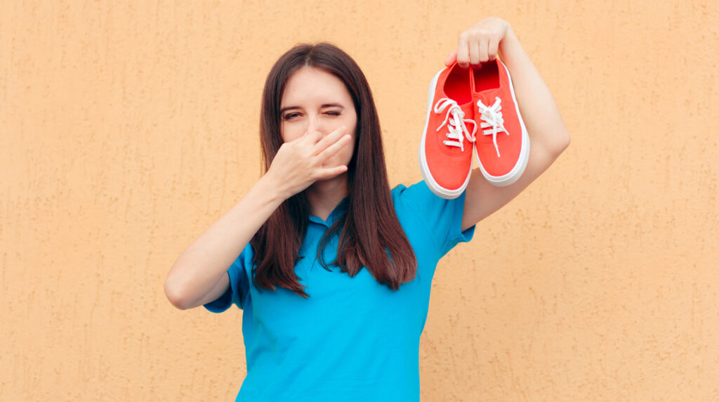 A woman covers her nose while holding up a pair of smelly shoes that need to be cleaned