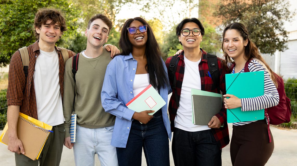 Five cheerful multiethnic college students standing together outdoors on campus, each holding notebooks and smiling at the camera.
